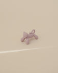 The mushie Cat Figurine Teether, a small purple cat-shaped baby teether with a curled tail, stands on a smooth beige surface under soft lighting.