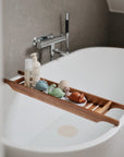 A modern white bathtub features a mushie Dino Mold Free Bath Play Set—colorful silicone bath toys with a wooden tray holding bottles and a bath bomb—in a minimalist bathroom with gray tiled walls.