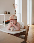 A baby in a beige-peach cap smiles in a high chair, playing with a toy attached by the mushie Adjustable Silicone Strap 2-Pack, surrounded by baby essentials in a bright, modern kitchen.