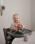 A smiling baby with a white bow sits in a high chair, holding mushie Chain Link Rings. A green bowl and pink cup are on the tray in a softly lit, minimalist room with flowers on the windowsill. Great for developing fine motor skills.
