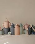 The mushie Silicone Puree Pouch and other pastel silicone baby feeding items, like cups, bowls, spoons, bottles, and pacifiers, are arranged neatly on a neutral background for mess-free feeding.