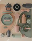 Mushie's Silicone Puree Pouch, along with pastel plates, bowls, cups, utensils, teethers, and a rocket toy, is displayed on peach and sage-green placemats for mess-free baby feeding in a stylish flat lay.