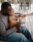 A man holds a baby on his lap while the baby reaches for a toy being handed by a woman. They are sitting together on a porch, and the baby is wearing a brown outfit, playing with a wooden toy.