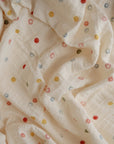 A close-up of the mushie Organic Cotton Muslin Swaddle Blanket with a crumpled texture, displaying small, colorful smiley face patterns in red, yellow, blue, and green scattered across the soft fabric.