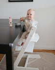 A baby in striped pajamas sits on a white high chair at a dark table, wearing a mushie Long Sleeve Bib, holding a spoon and facing the camera. A bowl and pink candle holder are on the table, with a striped mat under the chair.