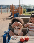 Two young children play with mushie Chain Link Rings on a bench at a playground, developing fine motor skills. Two apples are on the table, with outdoor play equipment and green grass in the background.
