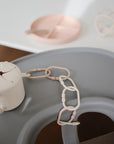 A mushie silicone snack cup, mushie Chain Link Rings in beige, and a divided pink plate with spoon rest on a gray high chair and white table in a softly lit room, ideal for developing fine motor skills.