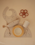 A beige baby placemat features a Mushie Classic Silicone Suction Plate, silicone bib, beaded pacifier clip, flower teether, sippy cup, yellow-rimmed bowl, and small spoon—perfect for mealtime with toddler plates that stay put.