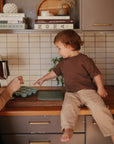 A young child sits on a kitchen counter, pointing at a Mushie Baby Food Freezer Tray while an adult pours liquid from a metal pitcher. Kitchen shelves, tiles, and books are visible in the background.