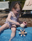Child by pool pours water on mushie Water Wheel Bath Toy in swimsuit.