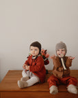 Two babies in cozy clothes and knit hats sit on a wooden table by flowers. One baby raises their hands, using the mushie Linen Pacifier Clip 2-Pack, while the other sits quietly with hands clasped.