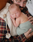 A bearded man in glasses holds a sleeping baby in knitted green pants, resting on mushie Organic Cotton Muslin Burp Cloths (2-pack) over his plaid shirt. A beaded pacifier clip hangs nearby.