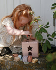 A young red-haired child sits outdoors on a stone step, fitting blocks into the mushie Shape Sorting Box in pastel pink. Framed by green leaves, this scene highlights hands-on learning and fine motor skill development.