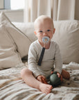 A baby sits on a beige couch, wearing a light onesie and holding a green toy, with sunlight streaming in, while using the FRIGG Little Viking Silicone 2-Pack pacifier from FRIGG.