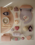 A neatly arranged collection of pastel mushie baby feeding items and toys, including a bib, utensils, plates, cups, a bottle, and the Princess Mold Free Bath Play Set on a light surface.