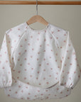 The mushie Long Sleeve Bib, featuring a wipe-clean, water-resistant design with small red bows, hangs on a wooden hanger against a plain beige background.
