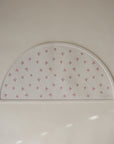 The mushie Silicone Placemat is a white, semicircular food-grade silicone mat featuring small red bow designs and the embossed mushie logo in the lower right corner, shown on a plain light surface.
