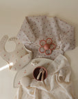 Flat lay of baby items in soft neutrals and cherry patterns, featuring the mushie Organic Cotton Muslin Swaddle Blanket along with a long-sleeve bib, flower teether, bowl and spoon set, textured ball, pacifier, and muslin cloth on a cream background.