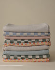 A neatly stacked pile of mushie's Organic Cotton Muslin Swaddle Blankets in various muted patterns and colors—stripes, solids, and florals—perfect as a baby swaddle or soft blanket, set against a neutral background.