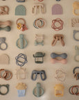 A display of 25 pastel-colored mushie Sun Teething Mitten silicone baby teethers, featuring animal, rainbow, circle, and geometric shapes. Neatly arranged on a light wall for a charming nursery accent.