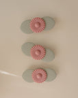 Three light green oval-shaped mushie Suction Spinner Toys with pink daisy-like decorations are arranged vertically on a beige surface, offering a delightful sensory experience.