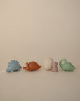 Four pastel dinosaur toys and a cracked egg from the mushie Dino Mold Free Bath Play Set are lined up on a beige background. These silicone toys in blue, orange, purple, and green ensure safe, mold-free bath time fun.