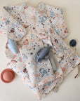 Two baby outfits with colorful space prints are displayed with a blue teether, pacifier clip, navy pacifier, red silicone bowl, and the mushie Water Resistant Wet Bag for easy diaper bag storage on a light background.