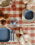 A table set for two with mushie Dinnerware Cutlery Set in blue, including dishwasher-safe plates and bowls on a red-and-white checkered cloth. Bread rolls, red juice, a blue flower teether, and napkins tied with red string add to the scene.