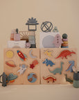 Mushie Wooden Puzzle, featuring alphabet pieces with dinosaur and space themes, sits on a light surface alongside pastel stacking toys and teethers—a playful collection that helps develop fine motor skills in a soft, neutral setting.