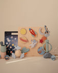 The Mushie Wooden Puzzle features pastel-colored, space-themed pieces like planets, a rocket, astronaut, and UFO to build fine motor skills—shown on a beige surface with a matching background.