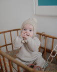 A baby in a beige knit outfit and pom-pom hat sits in a wooden crib, gazing up with wide blue eyes. mushie Chain Link Rings hang on the side, ideal for encouraging fine motor skills during playtime.