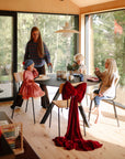 A woman serves breakfast to three children at a modern dining table in a sunlit room with large windows. mushie Dinnerware Cups are on the table, while a chair with a large red bow is in the foreground and greenery is visible outside.