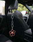 A mushie Chain Link Rings toy in a pink flower shape hangs from a car seat headrest inside a black car. The scene is bright, with greenery visible through the window.
