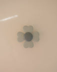 The mushie Suction Spinner Toy features a light gray, four-petaled flower design with a round center, made of silicone and mounted on a smooth beige surface, providing a calming sensory experience.