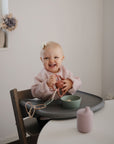 A smiling baby in a mushie Long Sleeve Bib and white bow sits in a high chair, holding a pink cup. A green bowl and pink bottle are on the table before her in a softly lit, minimalist room.