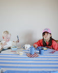 Two children sit at a blue-striped table with mushie Square Dinnerware Plates. The toddler eats with a spoon from the BPA-free plates, while the older child in a pink hat playfully enjoys spaghetti among assorted dishes and cups.