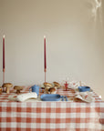 A table set with a red and white checkered tablecloth, mushie Square Dinnerware Plates in blue BPA-free plastic, utensils, a napkin tied with a bow, scattered mushrooms, and two tall red candles, all against a beige wall.