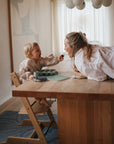 A young child feeds a smiling woman as they share a joyful moment at the table, with the Mushie Splat Mat protecting the floor in their cozy, modern, light-filled room.