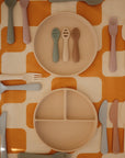 A Mushie Classic Silicone Suction Plate and a round non-toxic beige plate sit on an orange and white checked tablecloth, surrounded by colorful silicone forks, spoons, and knives.