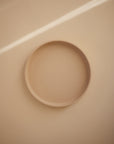 The Mushie Classic Silicone Suction Plate, round and beige, rests on a smooth light beige surface with soft lighting creating a diagonal highlight in the background.