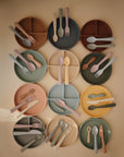 Twelve Mushie Classic Silicone Suction Plates in pastel shades, each with a matching fork, spoon, and knife, are arranged in a grid on a neutral surface, showcasing these non-toxic plates in a colorful, organized display.