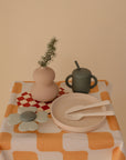 A beige table with a yellow and white checkered cloth holds a round vase with green foliage, the Mushie Classic Silicone Suction Plate with cutlery, a green sippy cup, a flower-shaped coaster, and a red and white napkin.