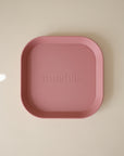 The mushie Square Dinnerware Plate is a pink, silicone, BPA-free baby plate with rounded edges and “mushie” embossed in the center, perfect for little ones’ meals. Shown on a light beige surface.