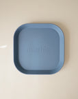 The mushie Square Dinnerware Plate is a matte blue, BPA-free plastic plate with rounded corners, raised edges, and "mushie" embossed in the center, shown on a light beige surface.