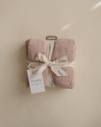 The mushie Organic Cotton Muslin Cloths 3-Pack in soft blush-pink, neatly folded with a cream ribbon and white tag, is perfect for nursery needs or your diaper bag, shown on a neutral beige background.