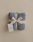 A neatly folded mushie Organic Cotton Muslin Cloths 3-Pack, tied with a cream ribbon, sits on a neutral background. A white tag reads “mushie”—perfect for nursery use or keeping handy in the diaper bag.