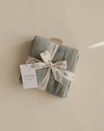 A folded light gray Organic Cotton Muslin Cloth from mushie, tied with a cream mushie ribbon and white tag, sits on a beige background—ideal for nursery use and easy to tuck in your diaper bag. Available in a handy 3-pack.