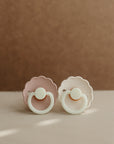 Two FRIGG Daisy Night Silicone Pacifiers, one pale pink and one white, with scalloped edges and round handles, rest side by side on a soft beige surface against a muted brown background.