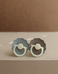 The FRIGG Daisy Night Silicone Pacifier 2-Pack by FRIGG, featuring scalloped edges in light blue and taupe, stands upright on a beige surface against a soft brown background.