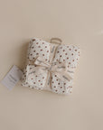 The mushie Organic Cotton Muslin Cloths 3-Pack, shown folded and tied with a branded ribbon, features white fabric with small red floral prints. Perfect for nurseries or diaper bags, each set displays a mushie tag on a beige background.